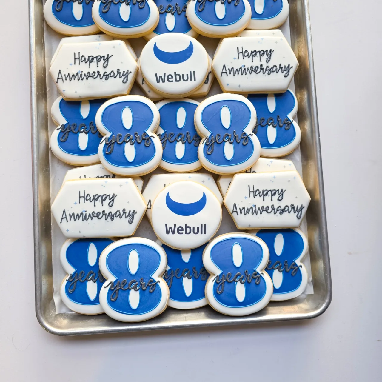 Blue, white, and charcoal Webull '8 years Happy Anniversary' corporate anniversary sugar cookies with printed Webull crescent logo rounds, blue number 8 cookies with 'years' script, and white hexagon 'Happy Anniversary' plaques with pale blue sparkle accents