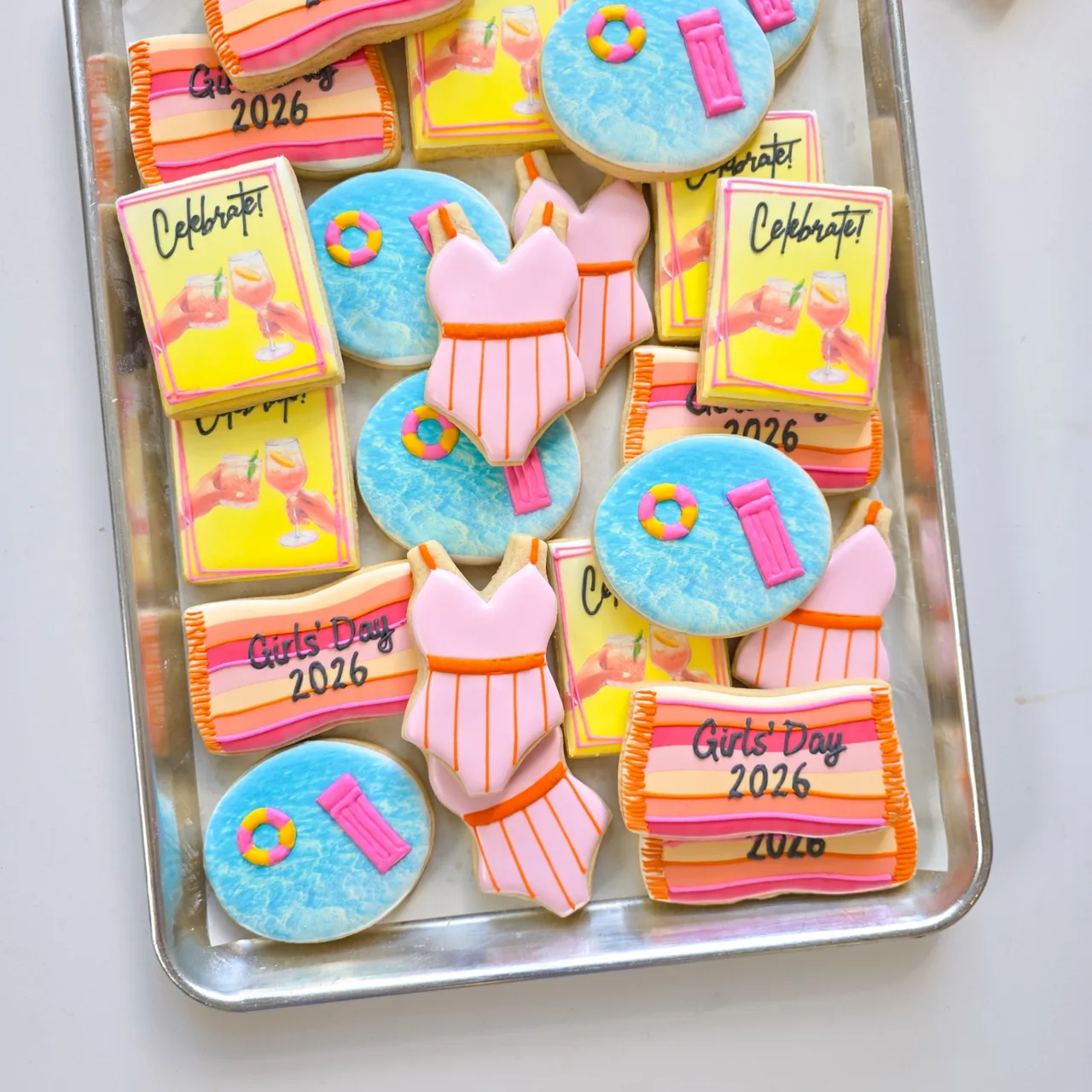 Pink, coral, orange, yellow, and blue 'Girls' Day 2026' pool party sugar cookies with striped beach towels, printed 'Celebrate!' cocktail cheers plaques, blue pool water rounds with pink floats and rings, and pink striped one-piece swimsuits