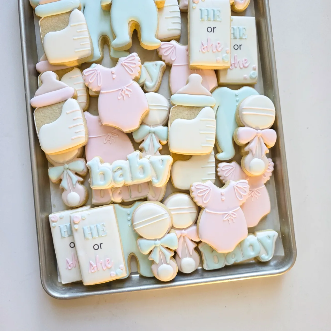 Pastel pink, mint, and cream 'He or She' gender reveal baby shower sugar cookies with 'baby' script plaques, pink ruffled dresses with bows, mint blue rompers, baby bottles, rattles with bows, and 'HE or she' rectangular plaques