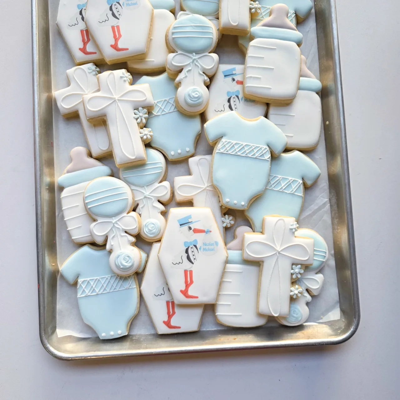 Soft blue and white 'Nicolas Michael' baby boy shower sugar cookies with printed stork hexagon plaques with 'Nicolas Michael' bundle, blue baby bottles, white and blue baby rattles with bows, blue baby onesies with lattice detail, and white christening crosses with floral accents