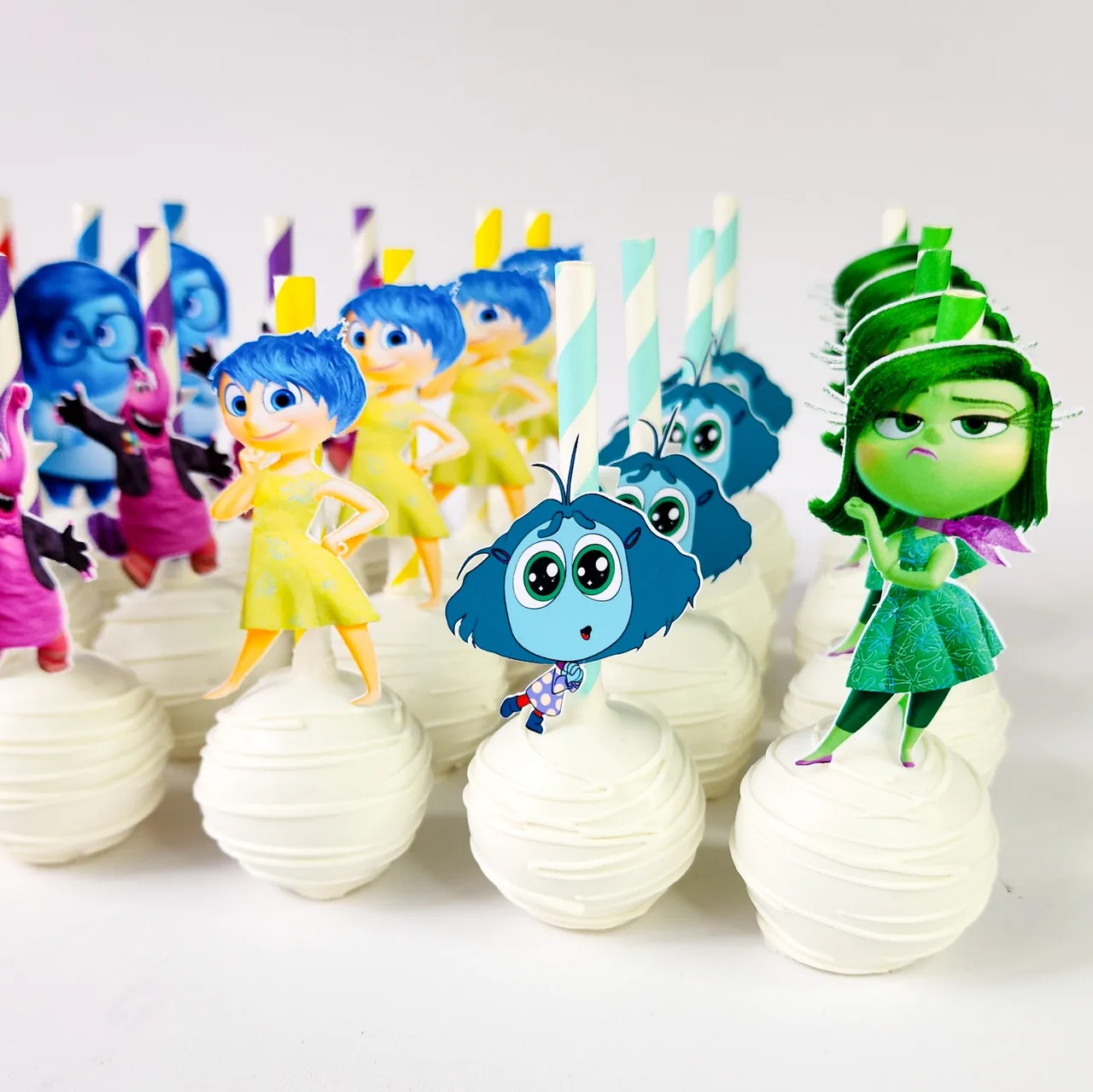 Inside Out 2 themed cake pops with white chocolate drizzle coating topped with edible printer character cutouts of Joy, Sadness, Anger, Envy, and Disgust on colorful paper straws