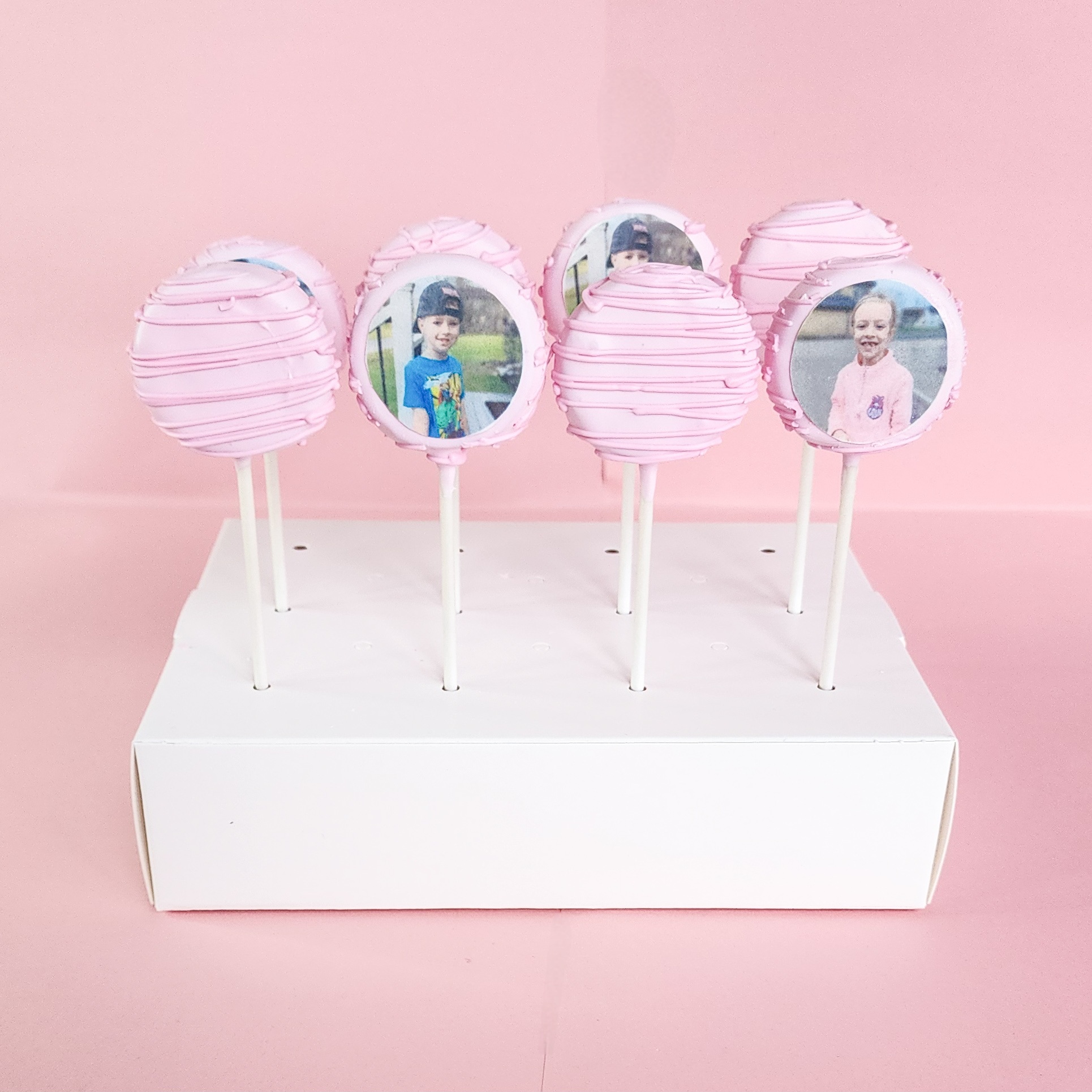 Pink drizzled cake pops with edible printer photo prints of children displayed on a white pop stand against a pink backdrop