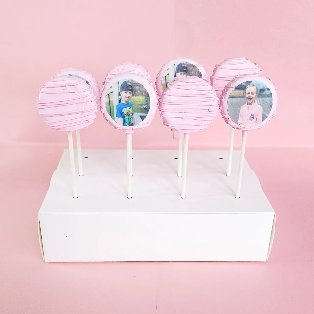 Pink drizzled cake pops with edible printer photo prints of children displayed on a white pop stand against a pink backdrop