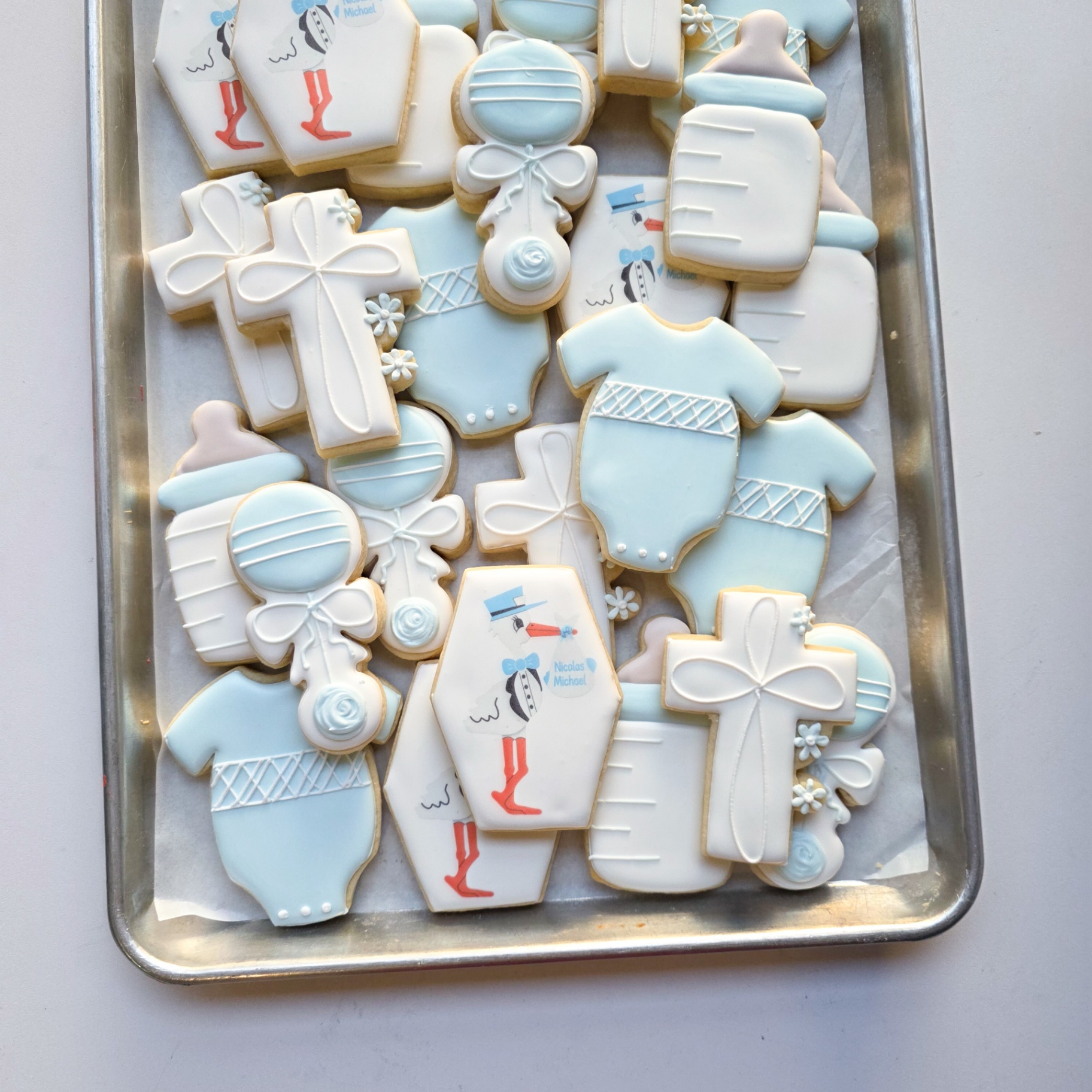 Soft blue and white 'Nicolas Michael' baby boy shower sugar cookies with printed stork hexagon plaques with 'Nicolas Michael' bundle, blue baby bottles, white and blue baby rattles with bows, blue baby onesies with lattice detail, and white christening crosses with floral accents
