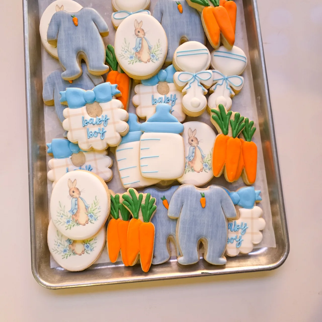 Dusty blue, white, orange, and green Peter Rabbit baby boy shower sugar cookies with printed Peter Rabbit oval portraits with blue floral wreaths, blue watercolor bunny onesies with carrot accents, orange carrot bunches with green tops, baby bottles, rattles, and gingham 'baby boy' bibs with blue bows