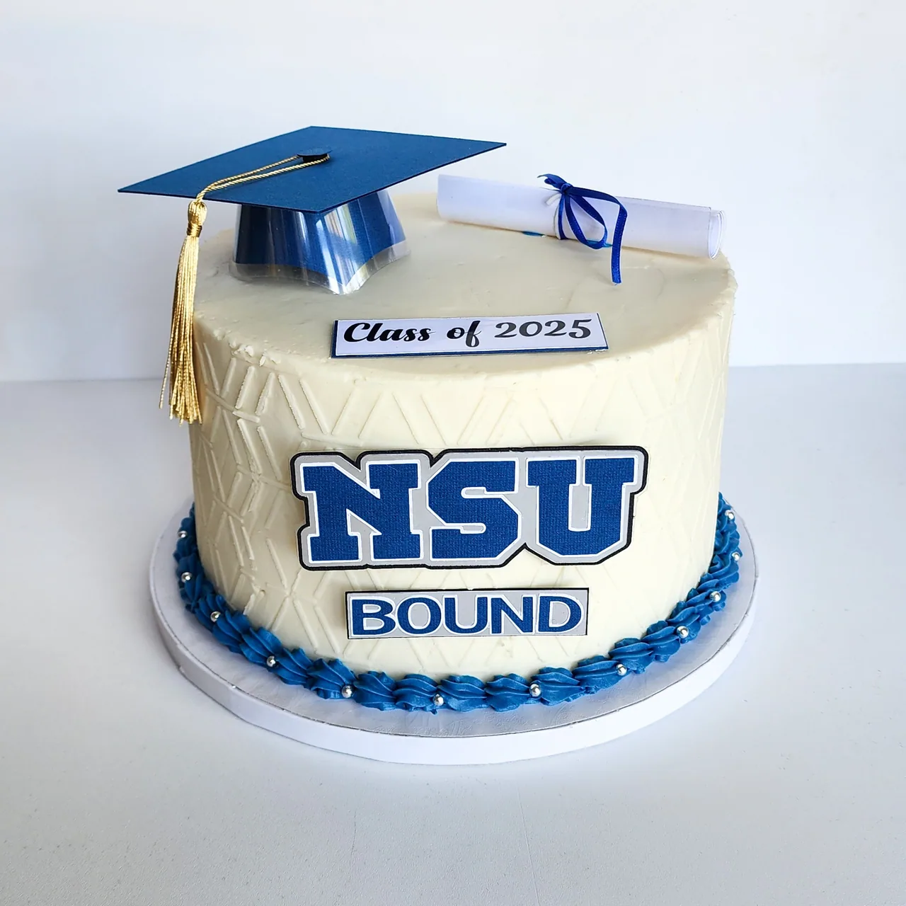Ivory graduation cake with NSU Bound topper, navy cap, diploma scroll, and blue piped border